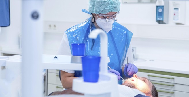 Dr. Josefine Holter in Schutzkleidung: Haube, Mund-Nasen-Schutz, blauem Kunststoffkittel und Handschuhen. Durch eine spezielle Schutzbrille mit Lupeneinsatz blickt sie in den geöffneten Mund des Patienten. Sie arbeiten mit feinen, metallischen Geräten. feinen 