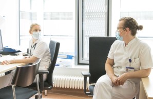 Dr. Louise Roggelin im Arbeitszimmer mit einer Kollegin, sie wenden sich einander zu. Beide in weißer Arbeitskleidung, sie tragen Mund-Nasenschutz-Masken. Die Haare sind bei beiden zum Zopf gebunden