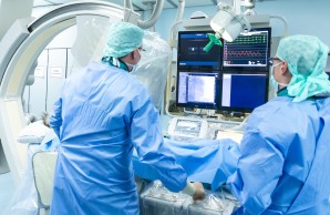 Dr. Marco Remmel im Katheter Labor bei der Arbeit, er und sein Kollege in blauer Schutzkleidung, sie blicken auf einen Monitor