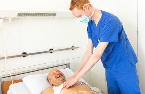 Niklas Ditsch, mit Mund-Nasen-Schutz und blauer Arbeitskleidung, beugt sich über einen im Bett liegenden Patienten. Dieser lacht herzlich