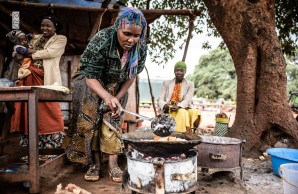 Eine Afrikanerin in traditioneller Kleidung, kocht draußen, über einen Kohlegrill gebeugt, neben ihr zwei weitere Frauen
