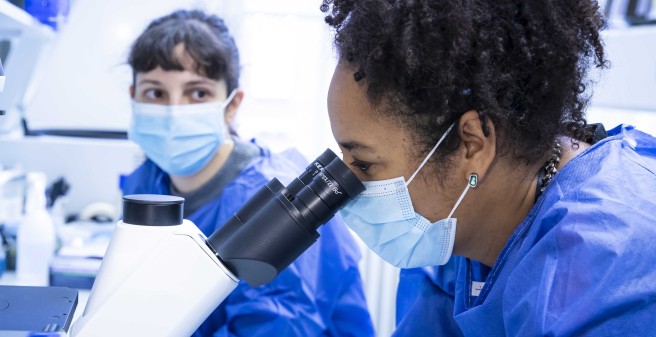 Prof. Dr. Marylyn Addo ihr Teammitglied Dr. Anahita Fathi im Labor. Frau Addo blickt in ein Mikroskop. Beide tragen blaue Arbeitskittel und Alltagsmasken