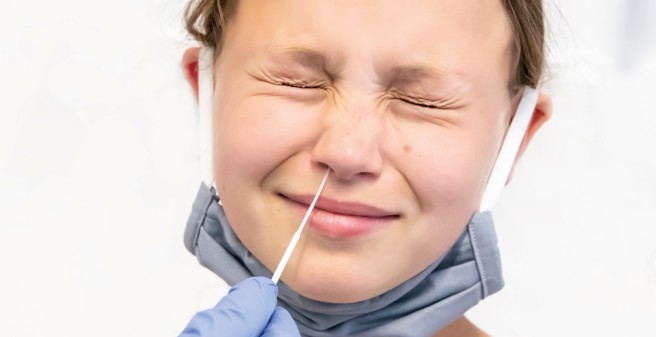 Nasenabstrich: ein dünnes Stäbchen wird in die Nasenhöhle eines Mädchens eingeführt. Die Jugendliche kneift die Augen zusammen.