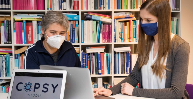 Priv.-Doz. Dr. Christiane Otto (l.) und Wissenschaftlerin Anne Kaman sitzen mit Mund-Nasen-Schutz an einem runden Tisch und blicken auf den Monitor eines Laptops. Im Hintergrund ein eng befülltes Bücherregal
