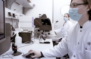 Die Mitarbeiter Nicola Wanner, im Hintergrund, und Prof Victor Puelles sitzen, in Kittel und Alltagsmaske, an ihrem Labor-Arbeitsplätzen. Vor ihnen jeweils ein Mikroskop und Monitor.