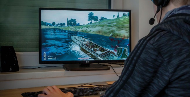 In einem abgedunkelten Zimmer sitzt ein Jugendlicher an der Tastatur seines Rechners. Er trägt Kopfhörer mit Mikrophon.Auf dem Monitor ist eine Spielsituation zu sehen: Kriegsschiff in einem Kanal, am Bildrand virtuelle Blutspritzer. 