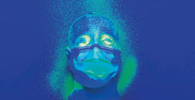 Maskentest. Auf einem blauen Untergrund liegt ein Dummy-Kopf mit einer Alltagsmaske. Grüne Partikel sind tröpfchenartig und großflächig neben und über dem Gesicht verstreut