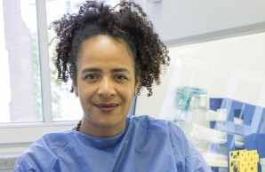 Prof. Dr. Marylyn  Addo blickt freundlich in die Kamera, sie sitzt neben dem Arbeitstisch, stützt lässig den Ellenbogen auf. Sie trägt eine blauen Schutzkittel und lila Handschuhe
